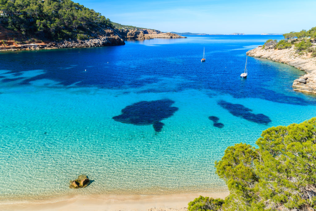 Luftaufnahme der Bucht von Cala Salada auf Ibiza mit kristallklarem Meer, umliegenden Grünflächen und einigen ankernden Booten