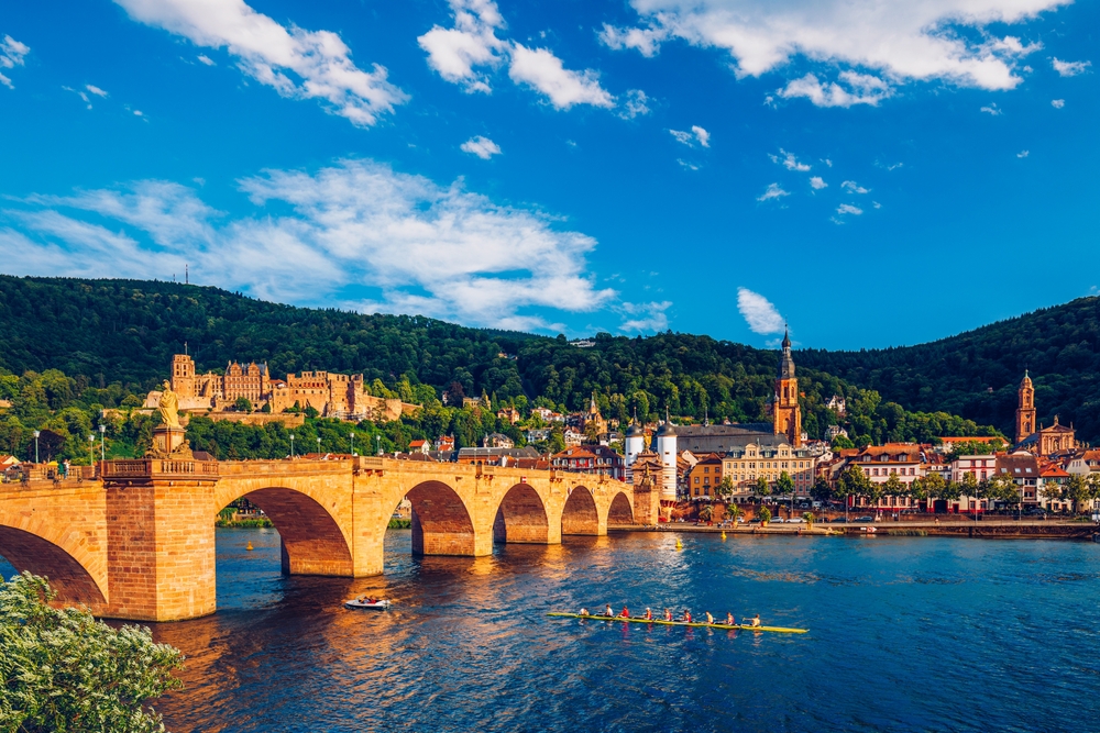 Heidelberg am Neckar mit einer von der Sonne angestrahlten Brücke und einem Ruderboot auf dem Neckar. 